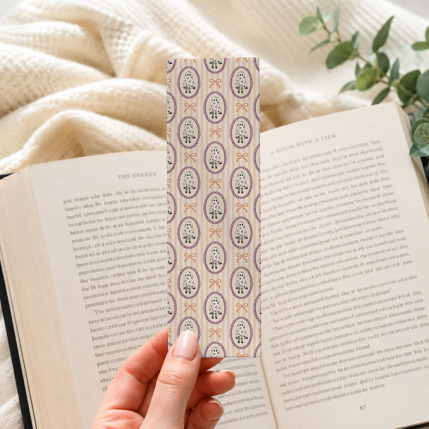 A hand holds a decorative bookmark with a repeating paisley pattern in muted purple, beige, and orange tones, positioned over open book pages in a cozy indoor setting with a cream knit blanket and green leaves in the background.