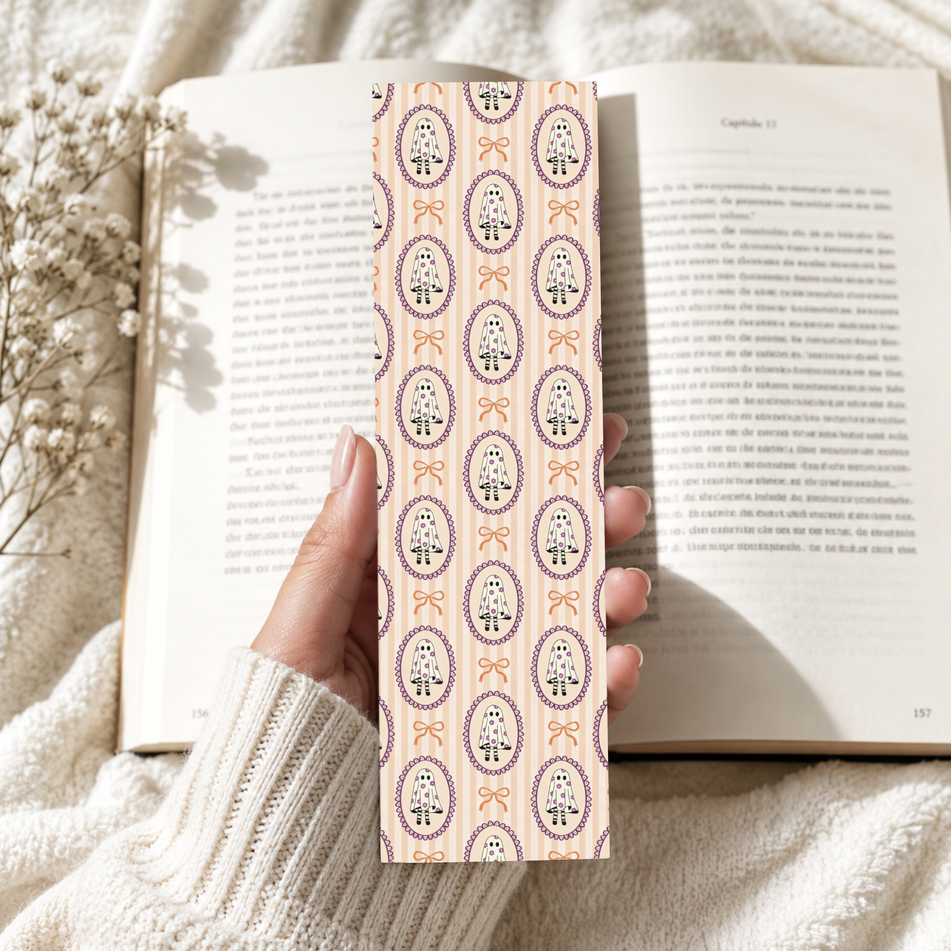 A hand holding a slim rectangular bookmark or card with a repeating ornate floral pattern in muted purple, peach, and cream, set against an open book on a soft cream knit blanket.