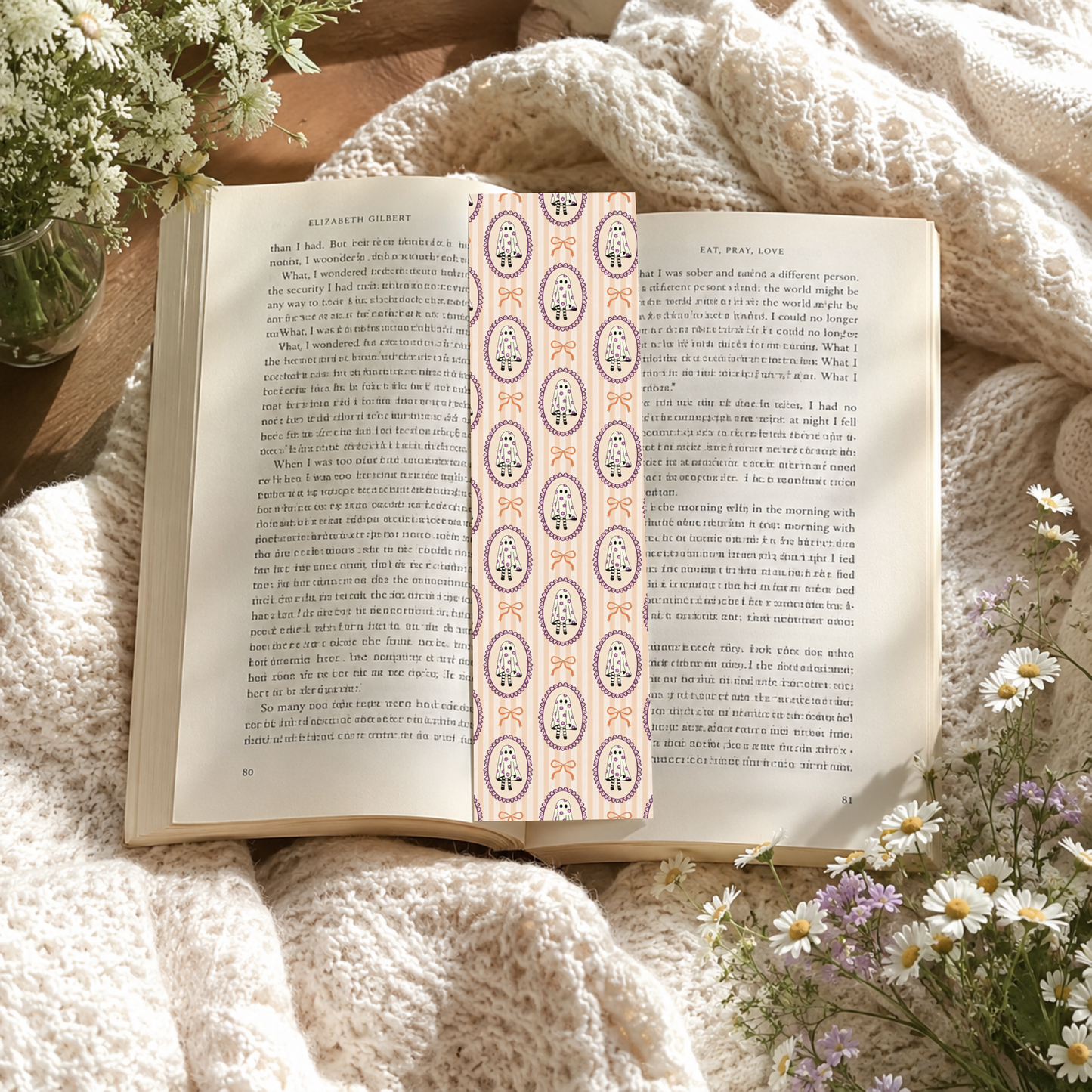 An open book spread on a soft cream knit blanket, decorated with white and purple daisies. A decorative paper bookmark with a vertical pattern of pink, purple, frames with cute ghosts and coquette bows is placed in the center of the pages.